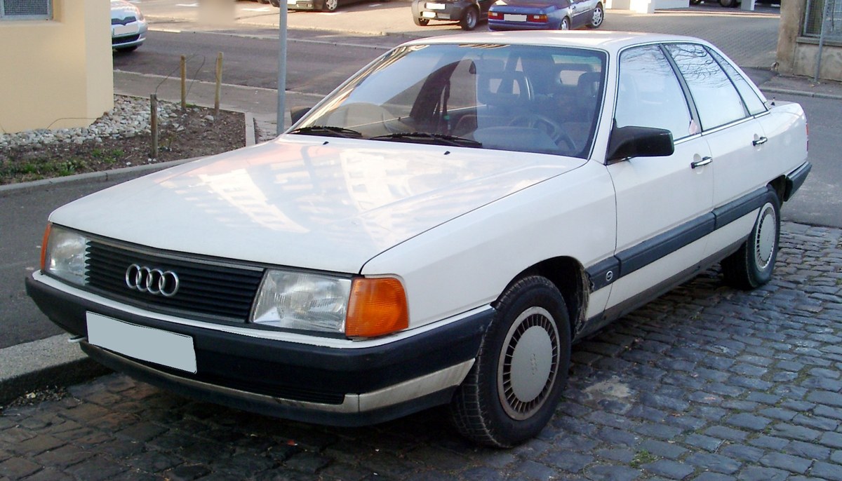 Белый Audi 100 C3 седан до рестайлинга, вид спереди слева в три четверти, на городской улице с брусчаткой