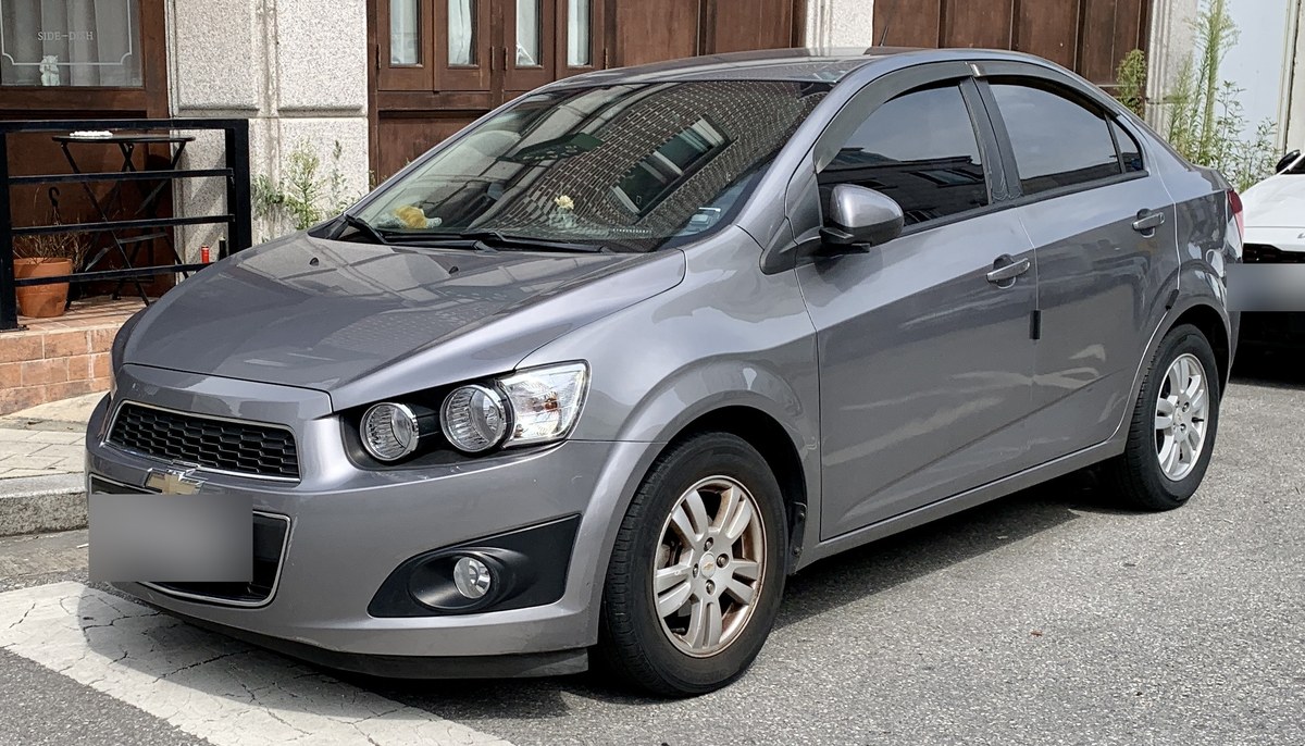 Серый седан Chevrolet Aveo T300 до рестайлинга, вид спереди сбоку, на городской парковке