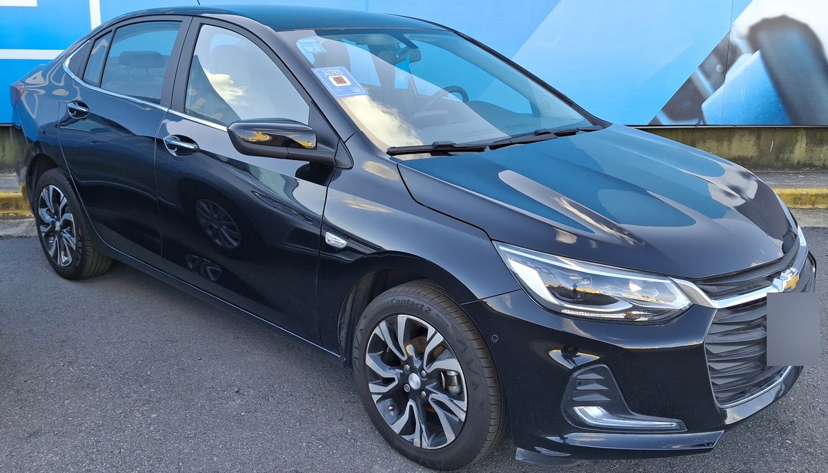 Чёрный седан Chevrolet Onix Plus, вид спереди сбоку, на парковке у рекламного щита