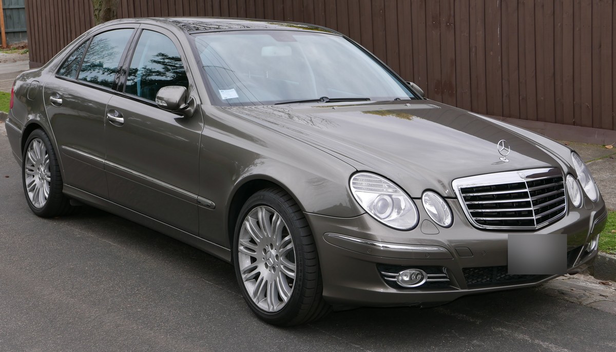 Коричневато-серый Mercedes-Benz E-Class W211 седан после рестайлинга, вид спереди справа в три четверти, у деревянного забора