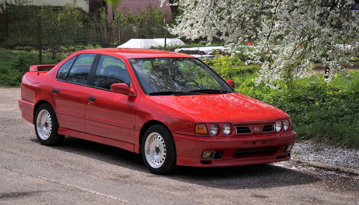 Красный Nissan Primera первого поколения (P10) eGT, седан, вид спереди в три четверти, на фоне цветущего сада
