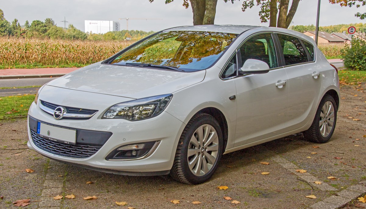 Белый Opel Astra J после рестайлинга, пятидверный хэтчбек, вид спереди сбоку, на парковке