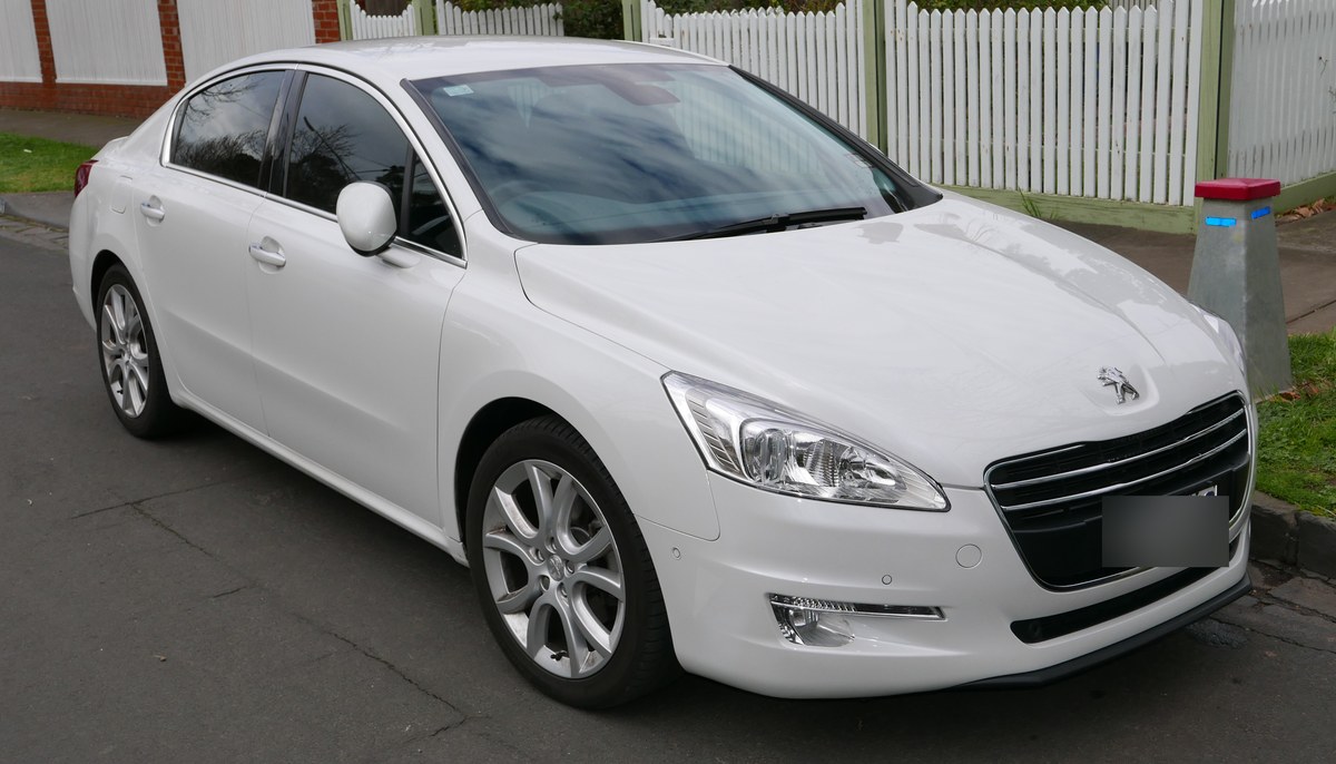 Белый седан Peugeot 508 первого поколения до рестайлинга, вид спереди справа, у здания