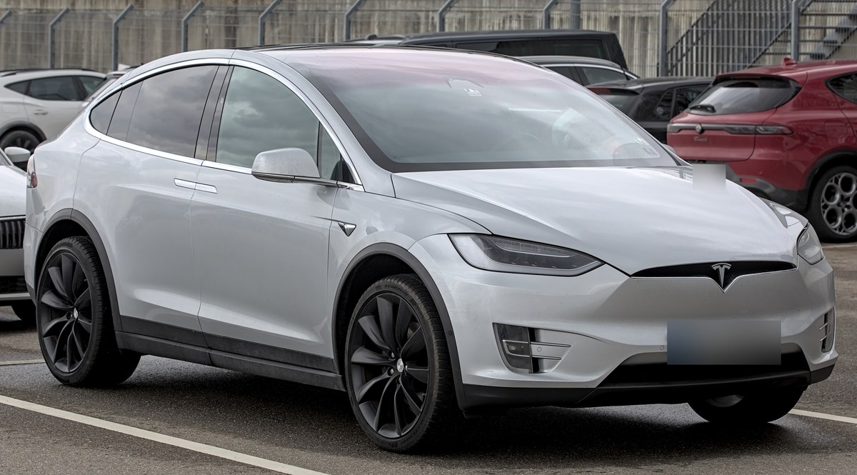 Серебристая Tesla Model X до обновления 2021 года, вид спереди в три четверти, на стоянке выставки