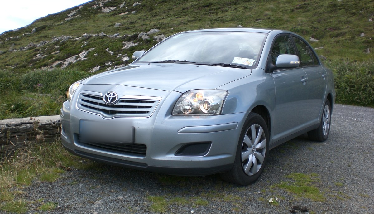 Серо-голубой Toyota Avensis T250 после рестайлинга, седан, вид спереди слева в три четверти, на гравийной площадке у склона
