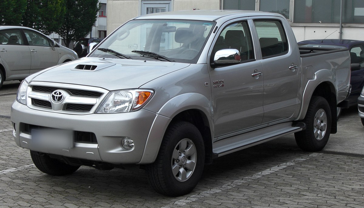 Серебристый Toyota Hilux седьмого поколения, пикап с двойной кабиной, вид спереди слева в три четверти, на парковке с брусчаткой